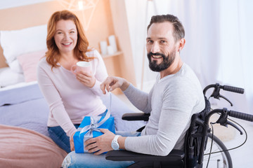 Fototapeta premium I am happy. Bearded disabled man smiling and opening a present while sitting in a wheelchair and a smiling woman sitting on the couch and holding a cup