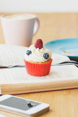 Small cupcake with raspberry and blueberry on the book, cup of hot cappuccino