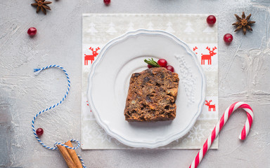 A piece of traditional Christmas English pudding (fruit cake) on white plate on a concrete light background. Christmas decor of the table. Funny napkins with reindeers Santa Claus.  Flat lay, top view