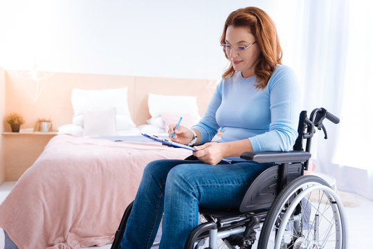 Doing job. Happy lovely smiling blond handicapped woman of middle age wearing glasses and writing while sitting in a wheelchair in a blue sweater and a bed in the background