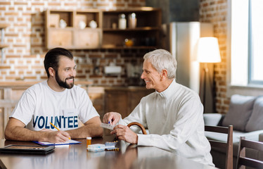 Lovely day. Cheerful pleasant aged man looking happy while sitting at the table and discussing his treatment with a kind attentive social worker