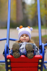 Baby girl in a funny hat swinging on the winter playground, perspective point of view. Looking at the camera