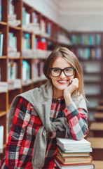 Portrait of a smiling casual student girl wears glasses holding stack of books in evening library, education concept