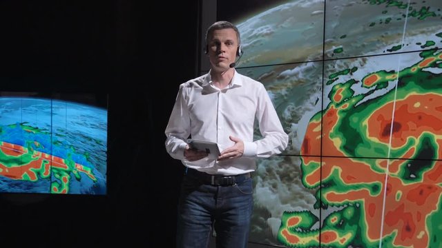 A news weather reporter wearing a headset presenting a live hurricane forecast displayed on two large screens. Elements of this image furnished by NASA.