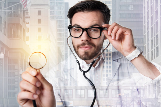 Curious doctor. Positive young handsome doctor looking curious while standing with a stethoscope and touching his glasses