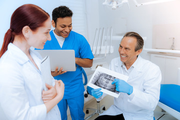 Fototapeta premium Professional diagnostics. Cheerful nice male dentist shoeing an X ray scan to his colleagues and smiling while pointing at it