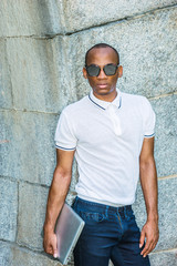 African American College Student studying in New York. Wearing white Polo shirt, blue jeans, carrying laptop computer, wearing sunglasses, black man standing by rocky wall on campus, looking forward..