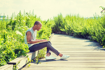 African American College Student studying in New York, wearing T shirt, black jeans, white sneakers, sunglasses, sitting on remote road with green tall grasses, working on laptop computer, smiling..