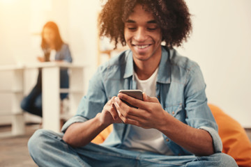 Cosy. Relaxed cheerful smiling student sitting in a comfortable orange beanbag chair and browsing the Internet in his smartphone
