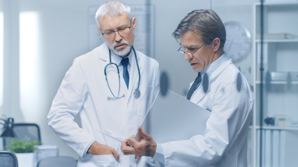 Two Specialist Doctors Discussing Patient's Log. Both are Senior and Experienced. Their Office Looks Modern and Respectable.