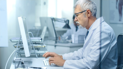 Fototapeta premium Senior Medical Practitioner Working in His Office at His Desktop Computer. Also His Assistant Works in the Background.