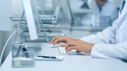 Close-up of Medical Practitioner's Hands Typing on a Keyboard Working on His Desktop Computer. In Background Assistant is Working.