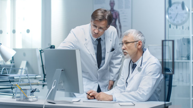 Senior Doctor and His Assistant Discuss Patient's Log on Personal Computer.