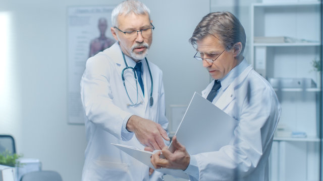 Two Specialist Doctors Discussing Patient's Log. Both are Senior and Experienced. Their Office Looks Modern and Respectable.