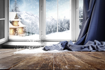 Wooden table with free space for your product or promotional text. Open window with snowfall. Landscape of winter forest mountains and small dock at the end of the village.