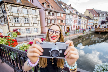 Portrait of a young woman tourist with photo camera standing on the old bridge in Colmar village in Alsace in north-eastern France - obrazy, fototapety, plakaty
