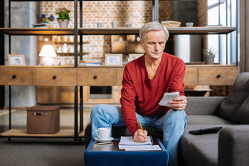 Financial problems. Serious pleasant retired man sitting at the coffee table and taking notes while counting his savings