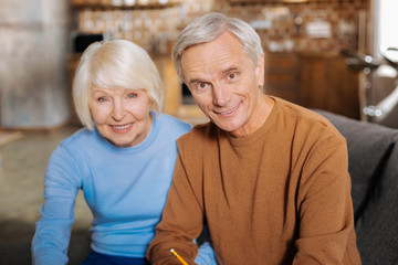 Elderly couple. Happy positive elderly couple sitting together and smiling while looking at you