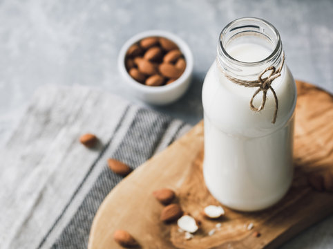 The almond milk in the glass bottle with almond nuts in the white bowl on the wooden decorative rustic cutting board.