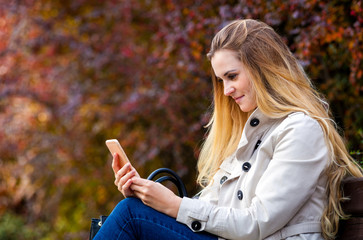Happy woman using smartphone sitting on bench at colorful autumn street