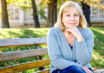   Middle-aged blond woman sitting on a  bench in the park looking at the camera on the autumn spring background. Portrait of beautiful senior happy elderly blond woman in a park on a sunny day.