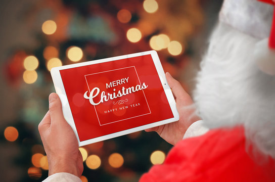 Santa holding tablet with Merry Christmas and a Happy New Year greeting. Bokeh and tree lights in background.