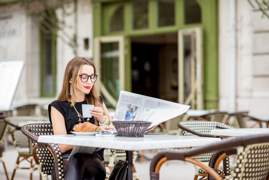 Young woman having a breakfast with coffee and croissant reading newspaper outdoors at the typical french cafe terrace in France