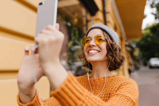 Spectacular young woman with happy smile making selfie with street on background. Cute girl wearing yellow sweater and sunglasses holding smartphone and taking photo of herself.