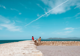 Young couple sitting on old city wall near ocean. Alghero. Sardinia. Italy.
