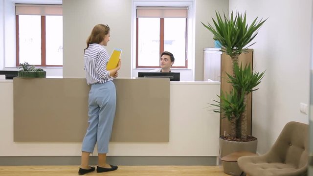 Woman at reception passes documents to man who sits talking on phone. Female employee passes folder with papers to male secretary, who is sitting at counter.