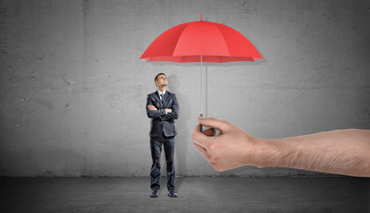 A small businessman stands looking up on a red open umbrella offered by a giant male hand.