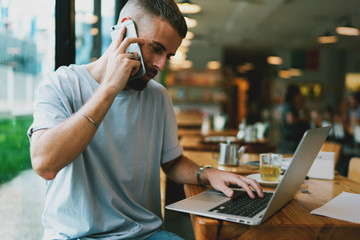 Handsome young man having conversation on a mobile phone while working remotely on laptop device. Bearded hipster guy is browsing websites on computer while talking by a smartphone.