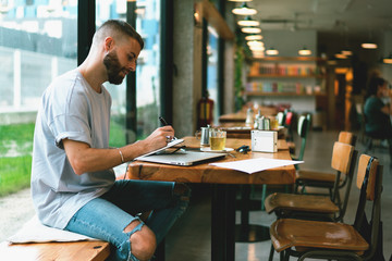 Handsome young designer is drawing graphic sketches for the project while sitting at wooden table in coffee shop interior. Thoughtful blogger is writing down new successful ideas for his blog.