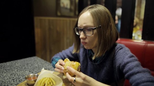 Young girl is eating untasty burger and French fries in the cafe. A woman in glasses is disappointed with the taste of stale cutlets in a hamburger and feel bad. Substandard food products, fast food.