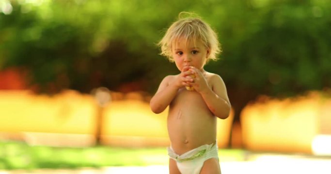 Candid shot of blonde infant toddler boy with diapers outdoors in 4k clip resolution. Toddler infant child holding a pear fruit outside