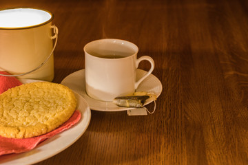 Hot steamy tea and cookie on a salmon colored napkin, white dish and wooden table with a candle