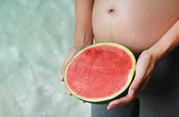 Pregnant woman sitting and holding cut half watermelon at her tummy.
