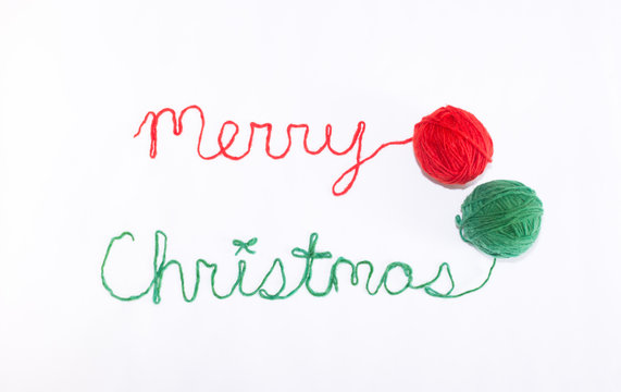 Merry Christmas Written in red and green yarn with Balls of Yarn on the Right. Photographed against a white background. 