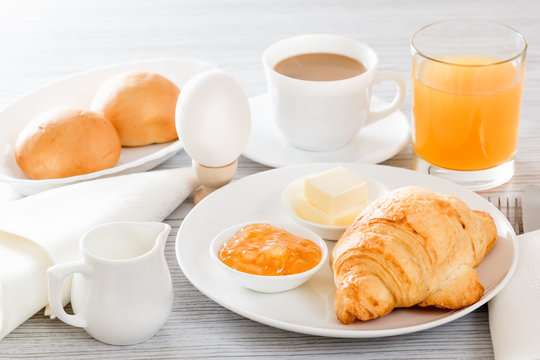 Continental breakfast with a croissant, boiled egg. Coffee or tea with milk, a glass of juice, buns, butter, jam. Soft backlight