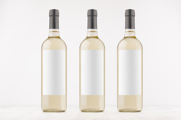 Three transparent wine bottles with blank white labels on white wooden board, mock up. Template for advertising, design, branding identity.