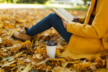 Woman in yellow coat, jeans and boots sitting under the maple tree with a red book and cup of...