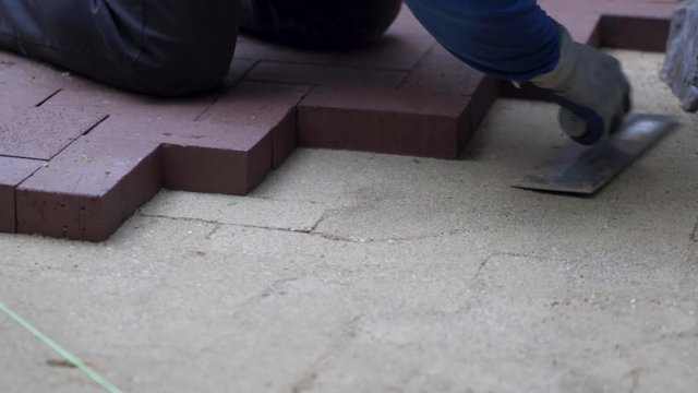 Closeup of a man smoothing out the sand preparing to lay down red brick pavers in a herringbone pattern.