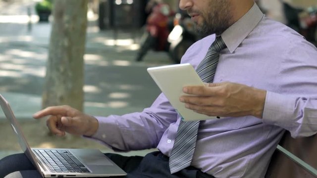 Young businessman with tablet and laptop talking to camera in city
