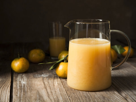 Pitcher and glass of citrus juice on rustic wooden background with fresh tangerines around.