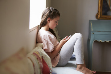 Smiling Girl Sitting On Window Seat Playing Game On Mobile Phone