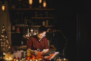 Beautiful girl in a red shirt packs Christmas presents on the eve of the new year in the kitchen with evening light.