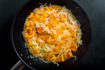 Chopped raw onion and fresh orange pepper in vegetable oil in a frying pan on a black background, closeup, top view