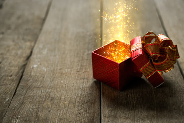 Red Christmas gift box with gold magic lights on wooden desk.