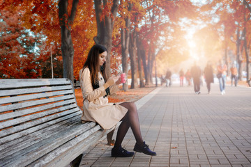 Fototapeta na wymiar Beautiful girl holding cellphone and coffee sitting on the bench at city autumn park