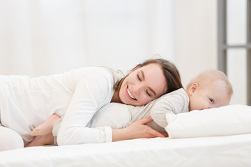 Happy cheerful young mom smiling playing with her little baby boy lying in bed at home. family portrait, white clothes, light house interior